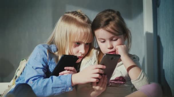 Дві дівчини встановлюють нові програми на своїх смартфонах, активно обговорюючи нові предмети. Сідайте на ліжко в моїй спальні — стокове відео