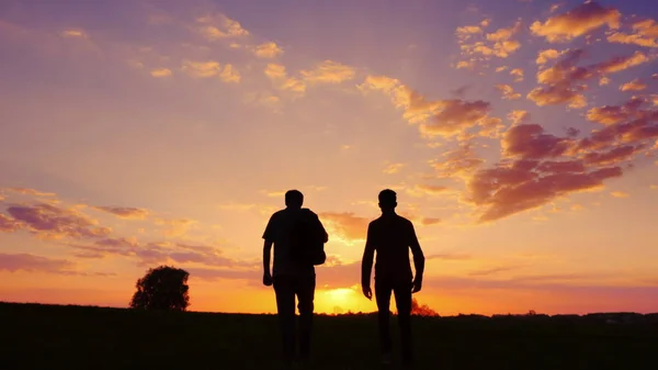 Siluety dvou mužů - otec a syn jdou ruku v ruce vstříc západu slunce. Pohled zezadu. — Stock fotografie