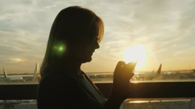Bir kadın bir akıllı telefon, hava alanında büyük bir pencere kullanır. Gün batımında, içinden uçakları görebilirsiniz
