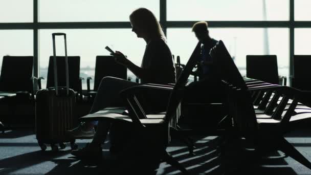 Siluetas de gente en el gran aeropuerto. Se sientan en anticipación del vuelo, utilizan el dispositivo: teléfonos inteligentes y tabletas — Vídeo de stock