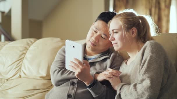 A young couple is resting at home, watching a video together on a tablet. Sit next to a white couch. Cozy house concept — ストック動画