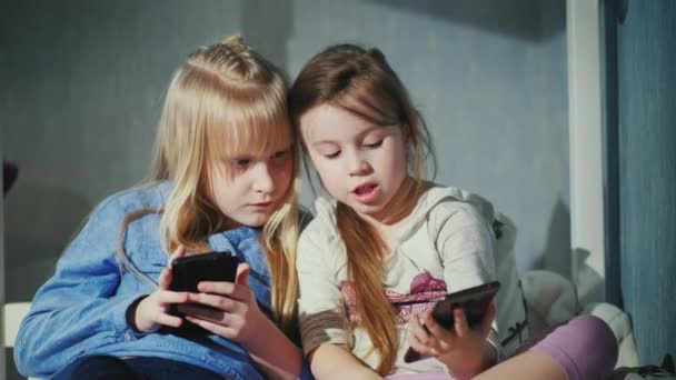 Свободное время современных детей. Двое детей играют со своими смартфонами, они сидят на кровати в собственной спальне — стоковое видео