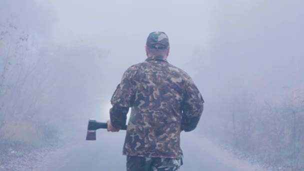 斧を持つ積極的な男が車に向かって霧の中を歩いている — ストック動画
