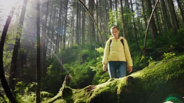 Kobieta podróżująca w kurtce i z plecakiem zawiązuje sznurek na butach. Stoi w lesie w malowniczym miejscu, promienie słońca pięknie oświetlają — Wideo stockowe