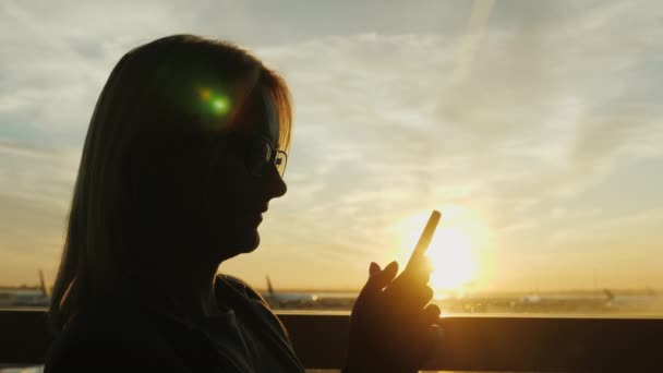 Silhouette im Profil einer Frau, die das Telefon in der Nähe des Terminalfensters am Flughafen benutzt. bei Sonnenuntergang — Stockvideo