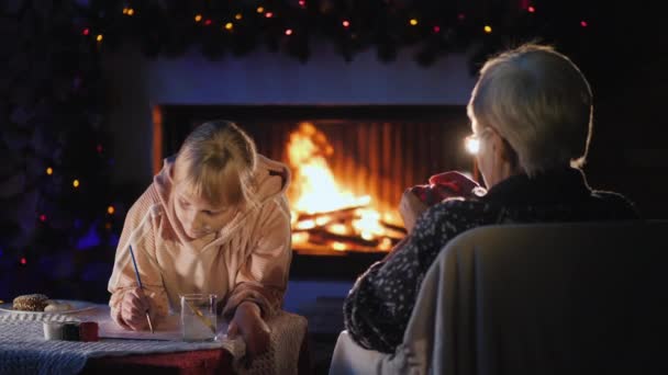 Randmother con una niña en Nochebuena descansar junto a la chimenea, la niña escribe una carta a Santa Claus — Vídeos de Stock