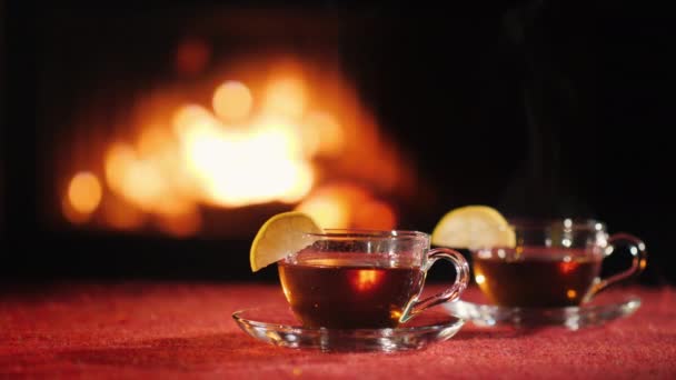 Dos tazas con té caliente en una mesa con un mantel rojo, en el fondo un fuego arde en la chimenea — Vídeos de Stock