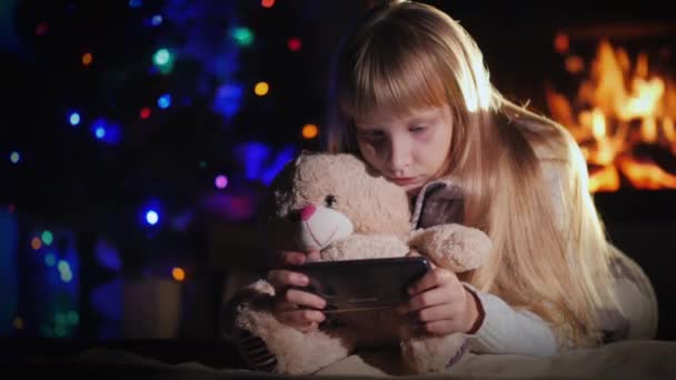 一个带着玩具熊的孩子在一个壁炉和圣诞树旁的智能手机上玩耍 — 图库视频影像