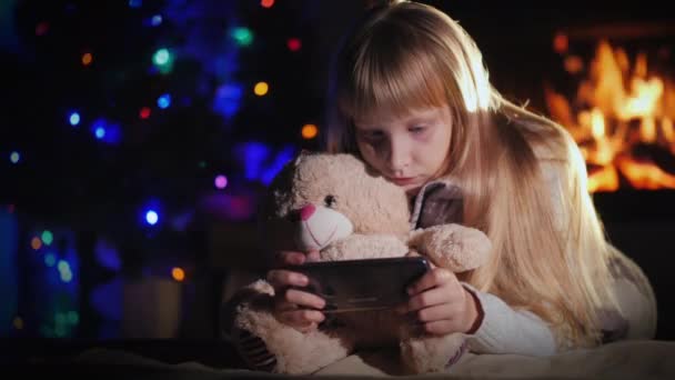 Seorang anak dengan boneka beruang sedang bermain di smartphone dengan latar belakang perapian dan pohon Natal — Stok Video