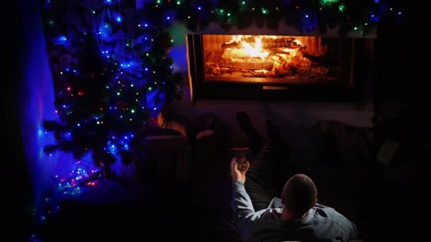 Ein Mann mit einem Glas Spirituosen sitzt am weihnachtlich dekorierten Kamin — Stockvideo