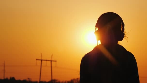 Silhouet van een tiener in de koptelefoon tegen de achtergrond van de stad en zonsondergang — Stockvideo