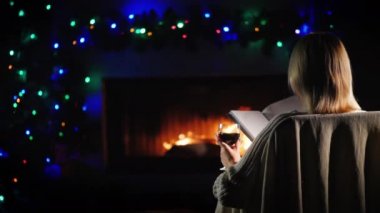A woman reads a book by the fireplace, next to the decorations for Christmas and New Year