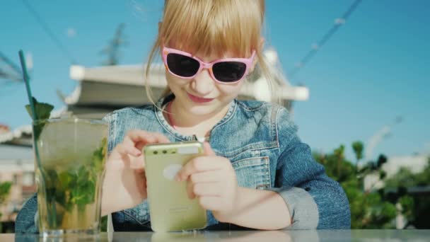 Chica divertida en gafas de color rosa juega en el teléfono inteligente. Se sienta en un apartamento de verano en una cafetería — Vídeo de stock