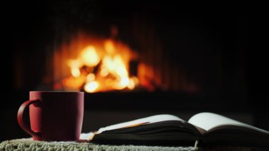 Şöminenin arka planında ateşin yandığı yerde açık bir kitap ve bir fincan sıcak çay.