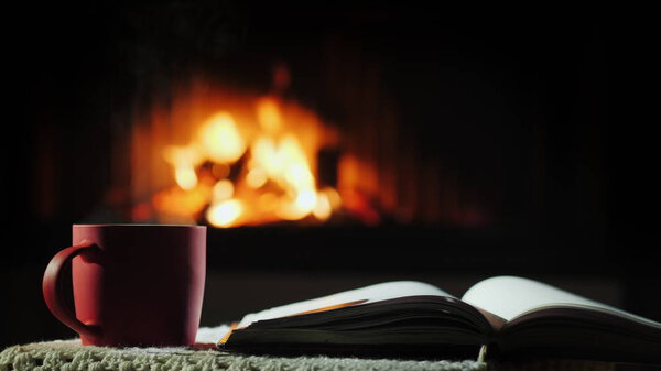 An open book and a cup of hot tea on the background of the fireplace where the fire is burning
