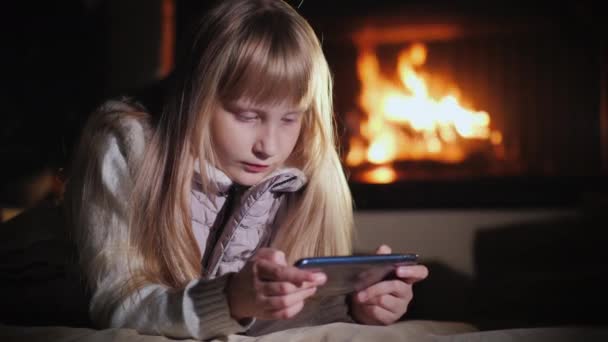Chica rubia utiliza un teléfono inteligente, se encuentra en el suelo de la habitación sobre el fondo de la chimenea — Vídeos de Stock