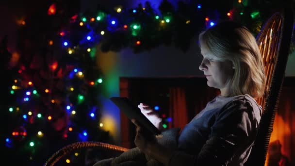 Vrouw gebruikt een tablet tegen de achtergrond van een open haard en feestelijke slingers — Stockvideo