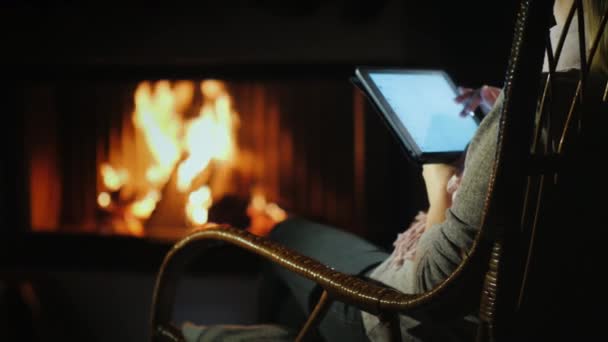 Een vrouw gebruikt een tablet terwijl ze in een schommelstoel bij de haard thuis zit — Stockvideo