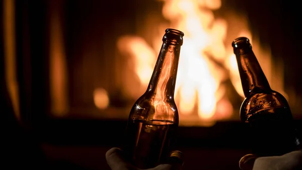 Dos hombres bebiendo cerveza junto a la chimenea - manos con botellas sobre un fondo de fuego ardiente — Foto de Stock