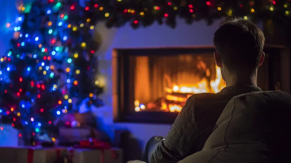 Un jeune homme admire le feu dans la cheminée, qui est décorée avec des guirlandes pour Noël. Vue arrière — Photo