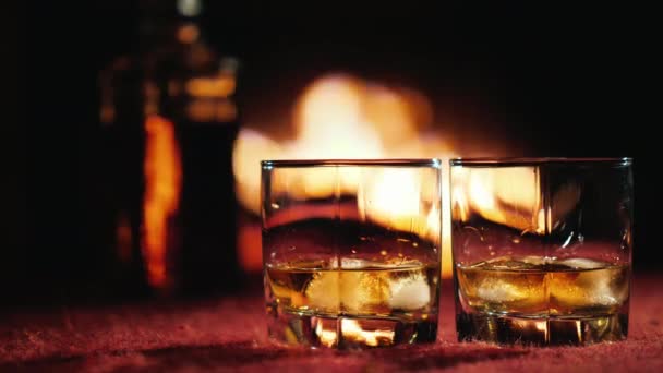 Два стакана виски со льдом, на заднем плане бутылка и камин, где горит огонь — стоковое видео