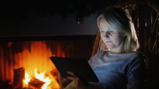 一位年轻女子在靠近火焰燃烧的壁炉的舒适的房子里使用数字平板电脑 — 图库视频影像