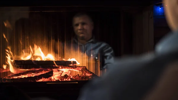 O fogo está a arder na lareira, um homem está a olhar para ele. Reflexão é visível no vidro — Fotografia de Stock