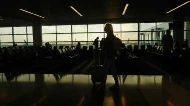 Takım elbiseli bir kadının silüeti. Bavuluyla havaalanı terminaline gidiyor. Batan güneş büyük bir pencerede parlıyor.