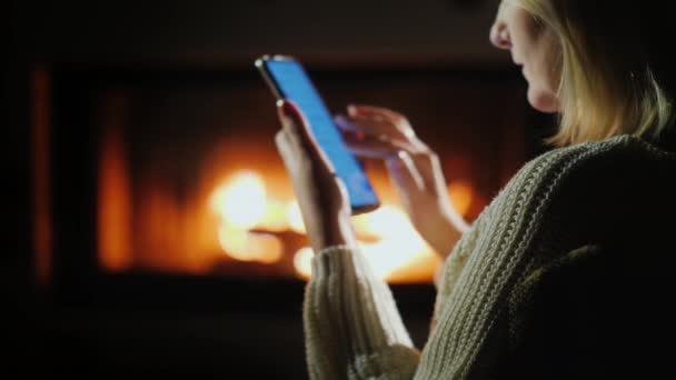 Mujer joven utiliza un teléfono inteligente mientras está sentado junto a la chimenea — Vídeos de Stock