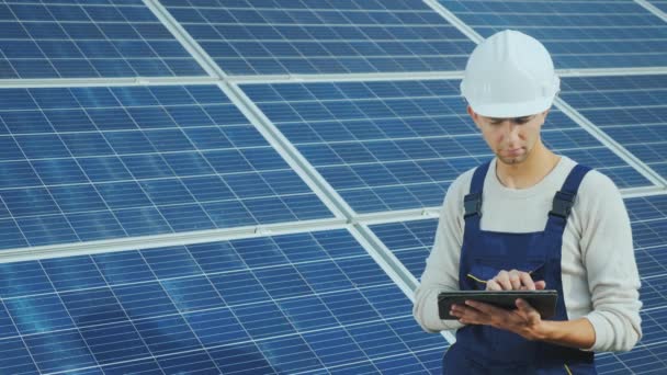 Jovem engenheiro em capacete branco trabalha com um tablet no fundo de painéis solares — Vídeo de Stock