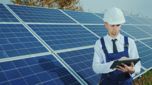 Een ingenieur in een helm en overall maakt gebruik van een tablet op de achtergrond van een zonne-energiecentrale — Stockvideo