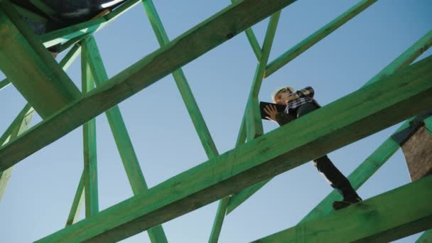 Constructeur féminin travaillant avec une tablette parmi les chevrons d'un toit inachevé — Video