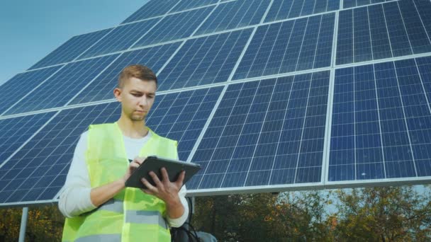 Un giovane ingegnere in abbigliamento da lavoro lavora con un tablet vicino a un grande pannello solare . — Video Stock