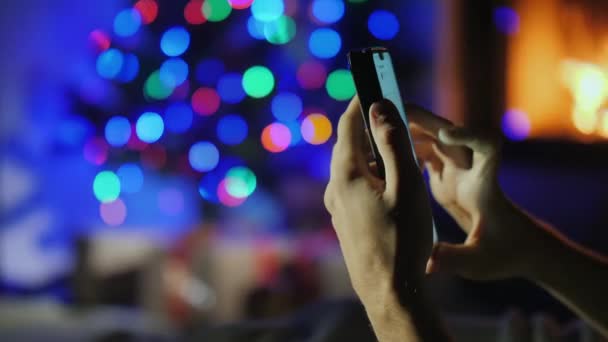 Manos con teléfono inteligente en luces borrosas del árbol de Navidad y la chimenea. Orden de regalo — Vídeo de stock