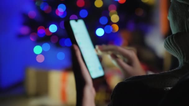 Vor dem Hintergrund der Weihnachtsbaumbeleuchtung hält er ein Smartphone in der Hand. Konzept der Geschenkbestellung — Stockvideo