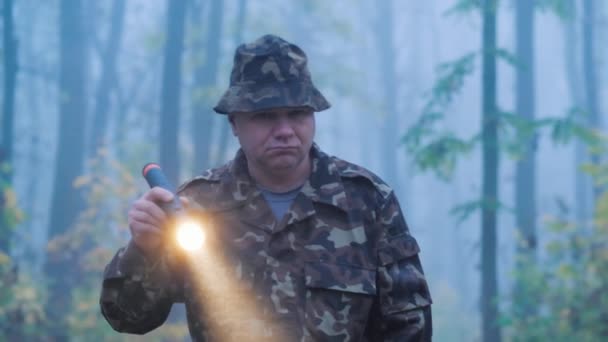 Egy álcázott férfi sétál az erdőben zseblámpával a kezében. Nedves esős idő kora reggel — Stock videók