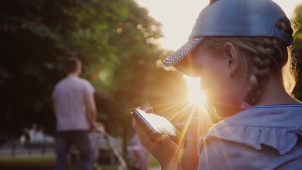 Ένα παιδί παίζει σε ένα smartphone, κάθεται σε ένα πάρκο σε ένα παγκάκι — Αρχείο Βίντεο