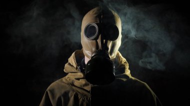 Koruyucu giysili ve gaz maskeli bir adam ön planda sigara içiyor.