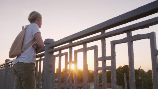 Mulher de meia-idade fica no corrimão da ponte, admirando o pôr do sol — Vídeo de Stock