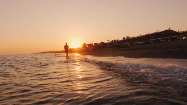 Silueta de un adolescente corriendo alrededor de la línea de surf al atardecer — Vídeo de stock