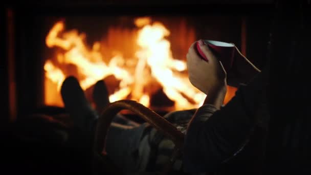 Ontspannen met een kopje ay bij de open haard, de handen van een vrouw met een rode beker zijn zichtbaar in het frame — Stockvideo