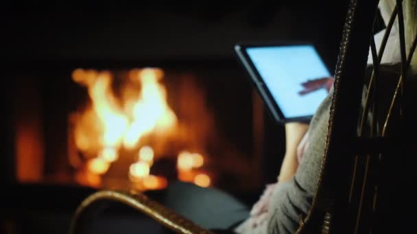 Rear view of A woman uses a tablet while sitting in a rocking chair by the fireplace at home — 비디오