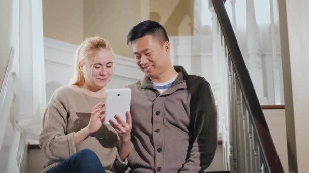 Mladý multietnický pár čte zprávy na tabletu. Sedí ve svém domě na schodech. — Stock video