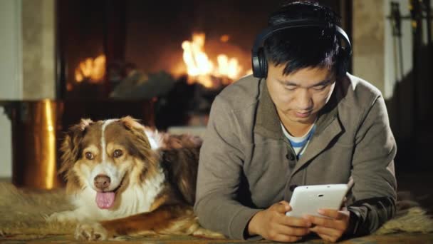 A young Asian man uses a tablet, resting at home near the fireplace. Nearby is his dog - Australian Shepherd — 비디오