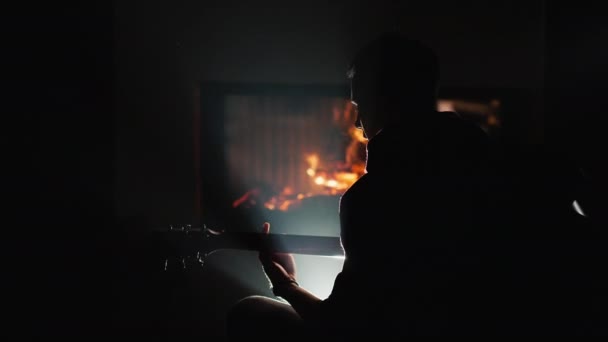Mladý muž hraje na kytaru, sedí sám u krbu — Stock video