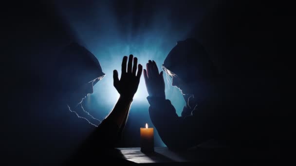 Двоє чоловіків в капюшонах моляться в темряві в променях світла — стокове відео