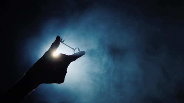 Een mensenhand houdt een sleutel vast tegen een donkere achtergrond waar mist kolkt. Zijaanzicht — Stockvideo