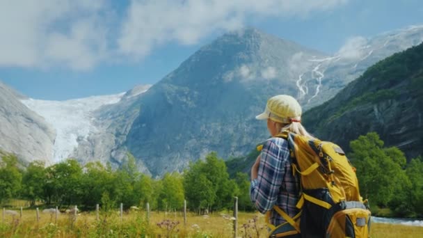 Una giovane donna con uno zaino cammina lungo la strada tra le montagne, viaggia attraverso i pittoreschi luoghi della Norvegia — Video Stock