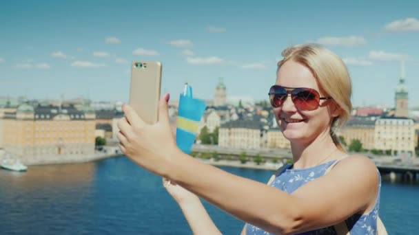 Mujer con la bandera de Suecia toma fotos de sí mismo contra el telón de fondo de la línea de la ciudad de Stockholms. Concepto Turismo en Escandinavia — Vídeo de stock