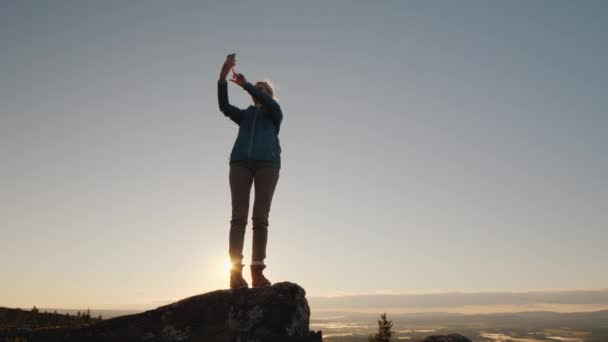 A traveler takes pictures of himself on top of a mountain at dawn — ストック動画
